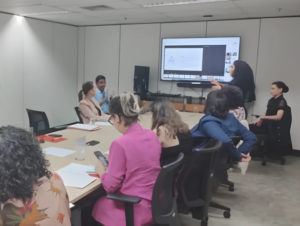 participantes da reunião sentados ao redor de uma mesmo, ao lado da mesa está uma monitor com a apresentação de slides. uma interprete de libras esta em pé sinalizando ao lado do monitor 