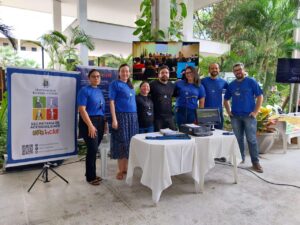 equipe ufc icnlui. 7 colaboradores da UFC inclui em pé stand na feira das profissões. ao fundo esta uma apresentação uma tv com um vídeo institucional. em primeiro plano temos duas messas com equipamentos de Tecnologia assistiva em cima