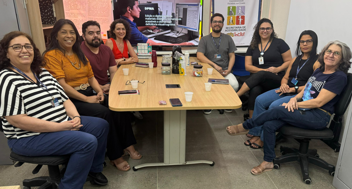 reunião com a vice-reitora da UFRS, ao redor de uma mesa estão as 8 pessoas sentadas que participaram da reunião. ao fundo está um banner com o logotipo da ufc inclui e um slide com um bolsista usando um scanner 