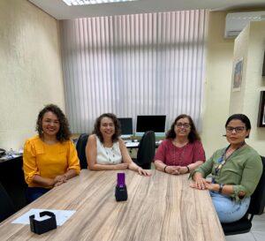 4 servidoras estão sentados ao redor de uma messa, em uma sala de reunião. no fundo estão 2 computadores