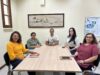 5 servidores, sendo 4 mulheres e um homem, estão sentados ao redor de uma messa, em uma sala de reunião. no fundo, na parede, está um quadro decorativo ao lado de um banner de um organograma