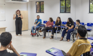 Reunião do fórum estudantil dentro uma sala de aula. onde um aluno sentado em uma cadeira de rodas fala ao microfone, sendo traduzido por uma interprete de libras ao seu lado.